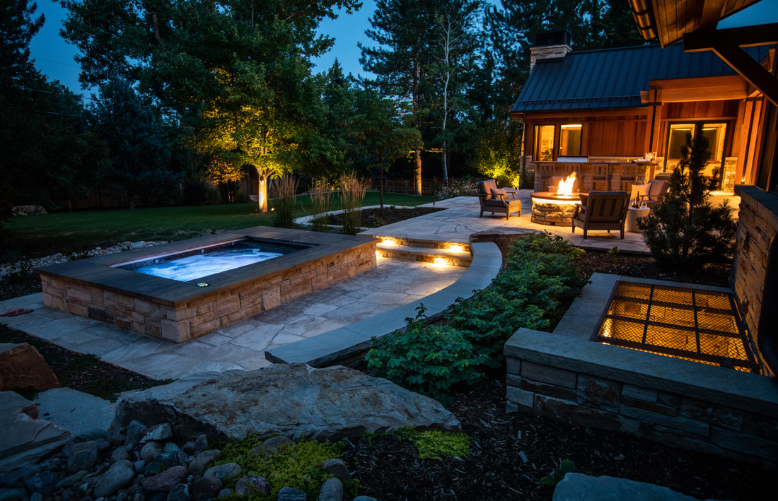 Native Edge outdoor living: hot tub, patio with fire feature, and stone hardscape at dusk